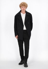 Abrigo negro de doble botonadura con un gran cuello, llevado sobre una camisa blanca de cuello en V y pantalones negros, combinado con botas negras de cordones.