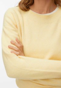 Suéter amarillo claro con textura, cuello acanalado y mangas largas, con un ajuste casual y tela suave. Los brazos están cruzados al frente.