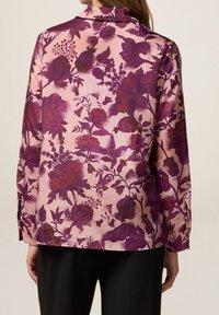 Floral patterned blouse in soft pink with deep purple flowers. Features a classic collar, long sleeves, and a straight hem. Smooth texture.