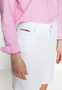 Jupe en jean blanc à coupe droite, dotée de poches latérales et de coutures multicolores sur les bords des poches. Associée à une chemise à manches longues rose clair.