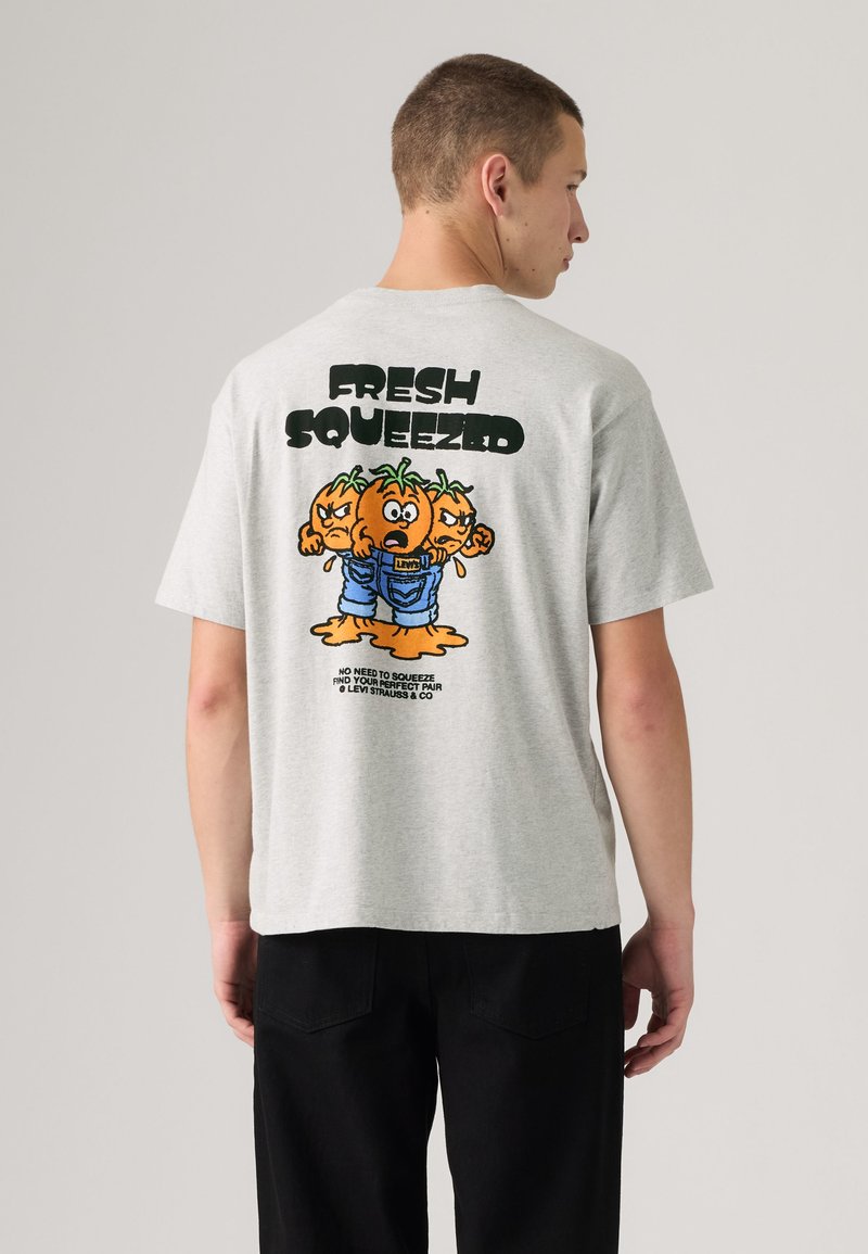 Junger Mann trägt ein hellgraues T-Shirt mit Cartoon-Darstellung von drei wütenden Tomaten in Jeans und dem Schriftzug „FRESH SQUEEZED“ auf dem Rücken.