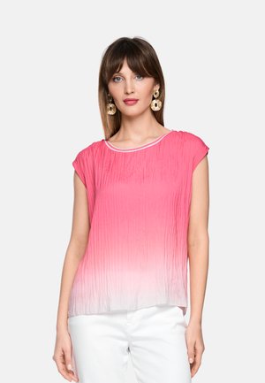 Frau trägt eine plissierte rosa-weiße Ombre-Bluse und weiße Hose, accessoriert mit großen goldenen kreisförmigen Ohrringen, steht vor einem schlichten Hintergrund.