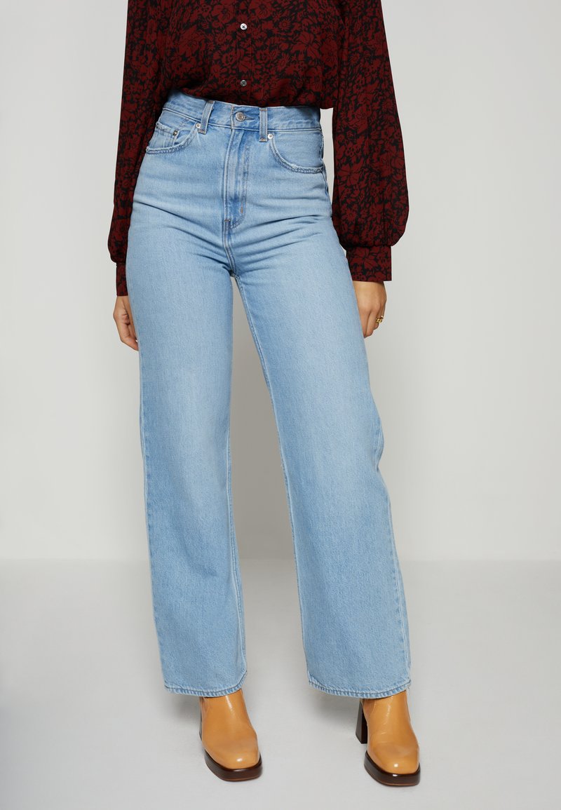 Jeans de cintura alta e perna larga em azul claro, com cinco bolsos, passantes para cinto e um pesponto discreto. Combinados com umas botas de cano curto castanhas de salto.
