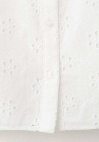 Tissu blanc avec broderie anglaise florale avec petites fleurs et feuilles, patte de boutonnage centrale avec deux boutons blancs visibles.