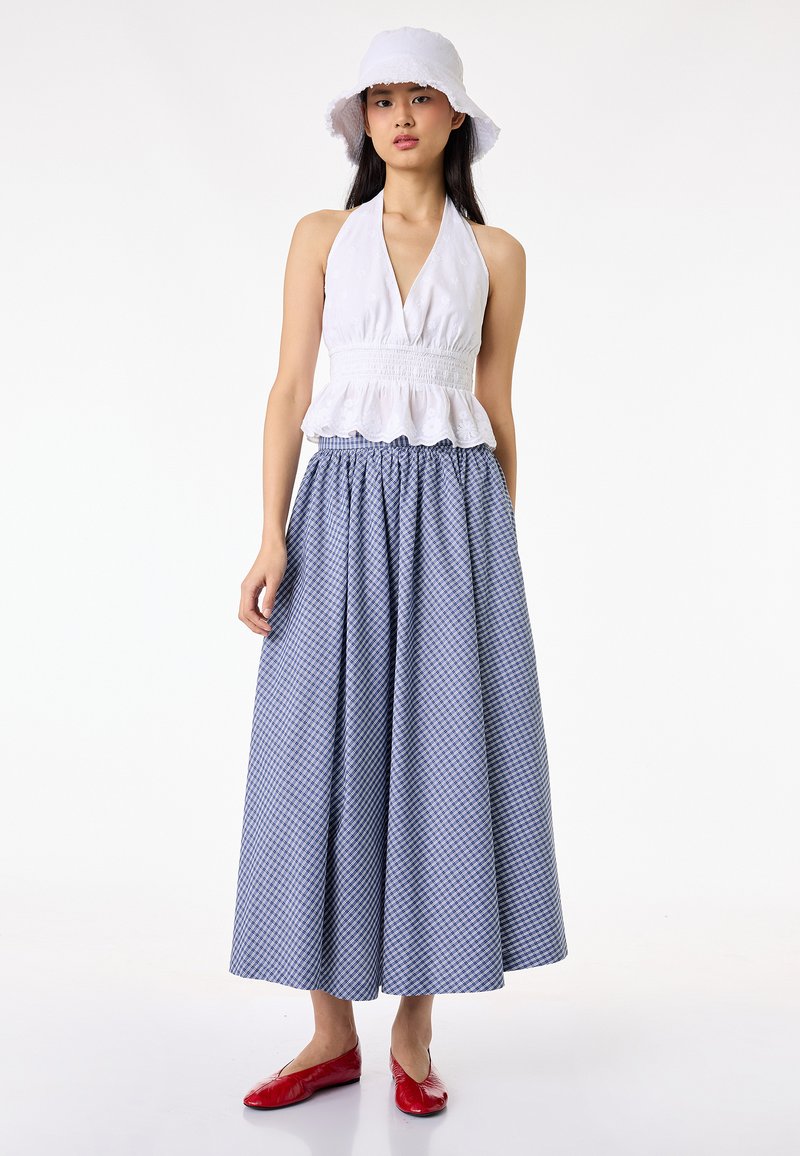 Femme portant un chapeau bob blanc, un débardeur blanc sans manches à col licou, un pantalon large à carreaux bleus et des chaussures plates rouges, debout devant un fond blanc.