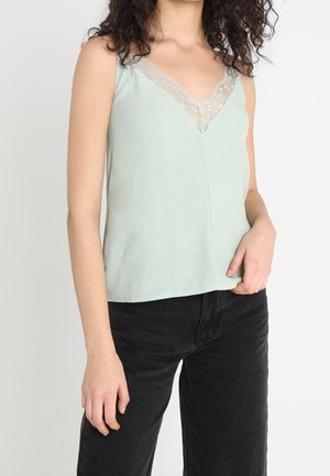 Kobieta ubrana w jasnooliwkowy bezrękawnik z koronkowym dekoltem w kształcie litery V oraz czarne spodnie, stojąca z jedną ręką przy talii.