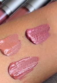 Tre nyanser av krämigt läppstift provade på medium hudton, med tre suddiga läppstiftstuber i bakgrunden.