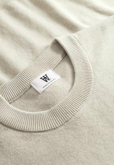 Pull gris clair avec un col rond côtelé. Texture douce, coutures visibles et une étiquette cousue affichant un logo stylisé.