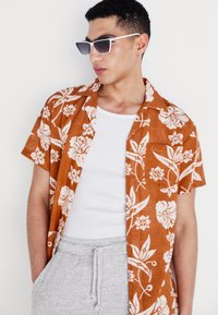 Camisa de botones de manga corta en color óxido con estampado floral blanco, combinada con una camiseta blanca sin mangas y pantalones de textura gris claro.