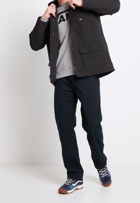 Schwarze Jacke mit mehreren Taschen über einem grauen Sweatshirt mit fettem Text. Dunkle Hose und Sneaker in Blau, Grau und Weiß runden das Outfit ab.