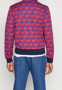Sweat-shirt à motif géométrique rouge et violet avec un col et des poignets côtelés bleu marine, porté par-dessus une couche blanche, accompagné d'un jean bleu.