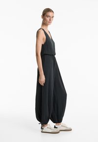 Črn kombinezon z rebrasto teksturo, globokim V-izrezom in širokimi hlačnicami, ki so zbrani pri gležnjih. Kombiniran z belimi supergami in rjavimi podplati.