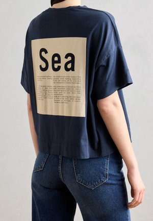 Persoon die een loszittend donkerblauw T-shirt draagt met een beige vierkant en grote tekst "Sea" op de achterkant, gecombineerd met blauwe spijkerbroeken.