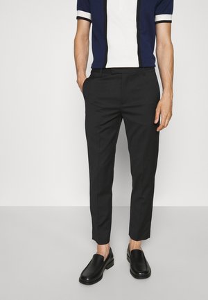 Uomo che indossa pantaloni neri aderenti, scarpe slip-on nere e una maglietta polo a maniche corte con righe verticali blu navy e bianche.