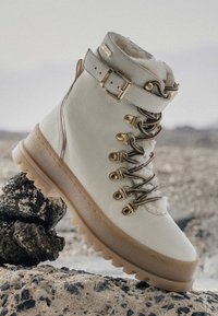 Botas de senderismo de cuero blanco con un cuello forrado de piel, herrajes dorados, cordones marrones y una suela de goma gruesa diseñada para ofrecer tracción.
