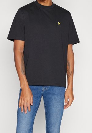 Mand iført en ensfarvet sort kortærmet t-shirt med en lille gul fuglelogo på brystet, kombineret med blå jeans, stående foran en hvid baggrund.