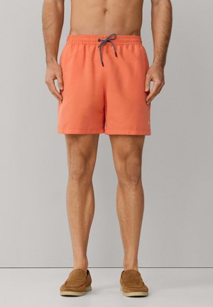 Man wearing coral swim shorts with black-and-white drawstring and brown slip-on shoes, standing against a gray background.