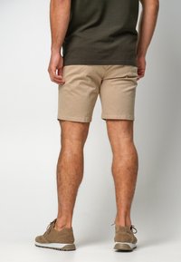 Shorts beiges en coton avec une texture lisse. La personne porte des chaussures en daim marron avec une semelle blanche, se tenant contre un fond neutre.
