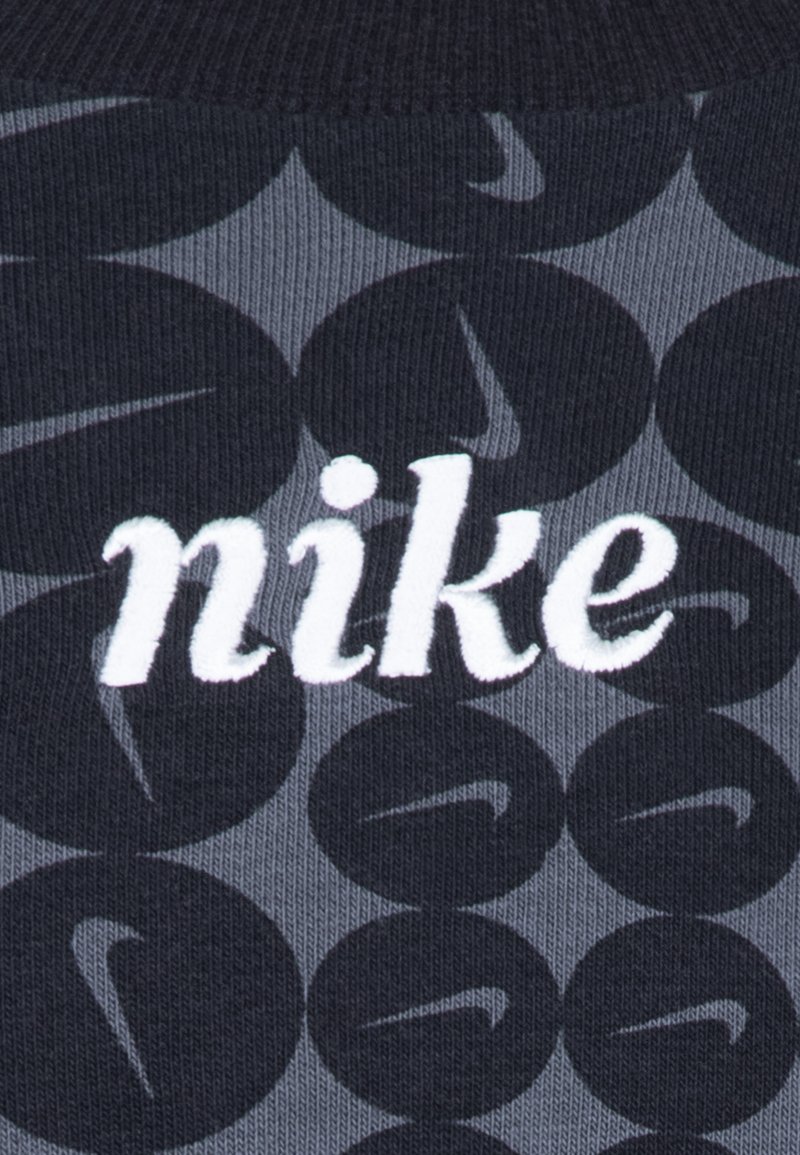Nike-logo op donkergrijze stof met een herhalend cirkelpatroon. De textuur is glad, met witte geborduurde letters.
