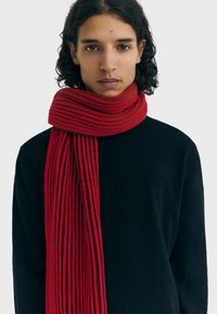 Young person with curly dark hair wears a black sweater and a red-and-black striped knitted scarf wrapped around the neck.
