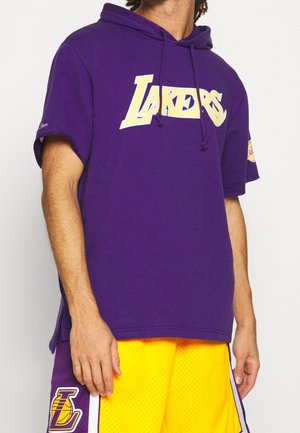 Persona que lleva una sudadera morada de los Lakers con letras amarillas y pantalones cortos de baloncesto a juego con el logo de los Lakers en el panel lateral morado.
