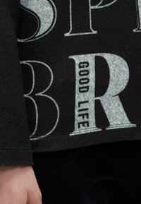 Schwarzer Pullover mit einem auffälligen, hellgrauen Grafikdesign, das den Text "GOOD LIFE" in großen stilisierten Buchstaben zeigt. Weiche Textur, lässiges Design.