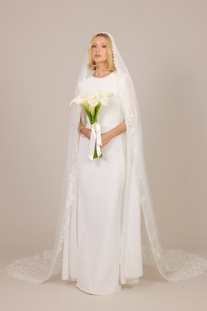 Panna młoda w długiej białej sukni i koronkowym welonie trzyma bukiet białych kielichów na jednolitym beżowym tle.