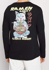 Zwarte longsleeve shirt met een afbeelding van een kat die ramen vasthoudt, met de teksten "RAMEN ADDICT" en "ALTIJD HONGERIG" in felle kleuren.