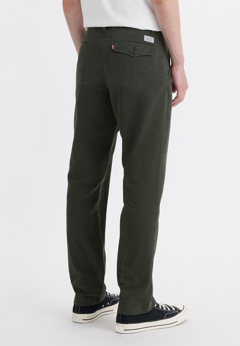 Pantalones de algodón verde oscuro con corte recto, bolsillos visibles y una etiqueta de marca. Llevados con zapatillas negras y blancas.