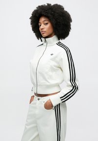 Fehér, rövidített fazonú, cipzáros dzseki magas gallérral, fekete csíkokkal az ujjakon és logóval a mellkason. Fehér nadrággal párosítva, oldalán zsebekkel.