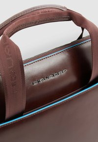 Bolso de cuero marrón con asa texturizada, logo plateado y detalle contrastante de cremallera azul. Presenta un diseño elegante y minimalista.