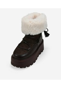 Botas de ante y cuero marrón con un puño de piel sintética blanca, puntera redondeada, suela gruesa color burdeos y cordones negros. Presenta un detalle de borlas en el talón.