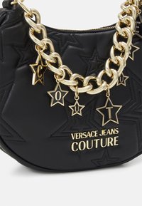 Černá kožená kabelka s texturou s reliéfními hvězdami, zlatým řetízkem a hvězdnými přívěsky. Vlastní logo "VERSACE JEANS COUTURE" v zlatém písmu.