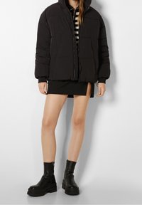 Svart pufferjacka med huva, med dragkedja framtill och quiltat mönster. Buren över en svart minikjol och kraftiga svarta ankelskor.