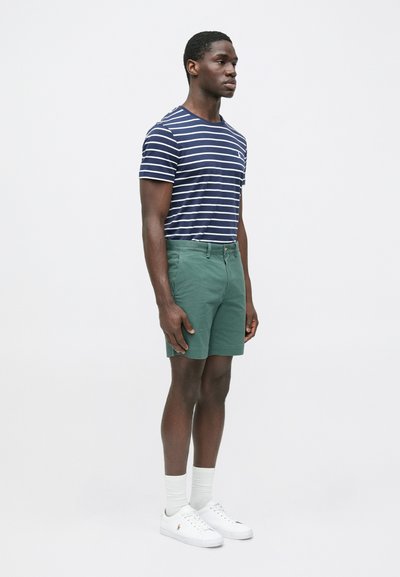 Μπλούζα με κοντά μανίκια σε ναυτικό μπλε και λευκές ρίγες, πράσινα σορτς, λευκά αθλητικά παπούτσια και ανοιχτόχρωμες κάλτσες. Απλός σχεδιασμός με χαλαρή εφαρμογή και ελάχιστες λεπτομέρειες.