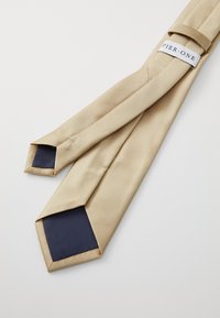 Beige silkeslöja med en slät textur, med en mörkblå inre foder och en etikett märkt "PIER ONE." Avsmalnande ändar och en subtil glans.