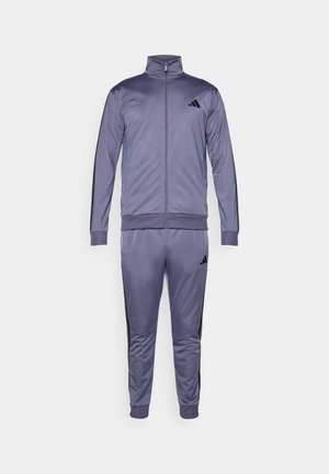 Fialový sportovní oblek obsahující zipovou bundu a kalhoty s rovnými nohavicemi, vyrobený z hladké látky s černými bočními pruhy a logem Adidas.