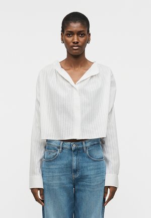 Γυναίκα που φοράει λευκό πουκάμισο με λεπτές ρίγες, κοντό με κουμπιά και φαρδιά γιακά, καθώς και ανοιχτό μπλε τζιν ψηλόμεσο, στέκεται μπροστά σε μια απλή φόντο.