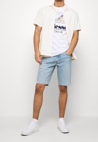Hombre vestido con una camisa de botones de manga corta blanca sobre una camiseta gráfica, pantalones cortos de mezclilla azul claro, zapatillas blancas, de pie con una mano en el bolsillo.