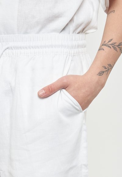 Indumento in lino bianco con cintura elastica e tasca laterale. Mano inserita nella tasca, che mostra una texture della pelle naturale e un tatuaggio.
