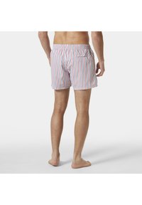 Hombre de pie descalzo, vistiendo un bañador de rayas verticales en blanco, rosa y azul, con cintura elástica y un único bolsillo trasero.