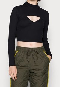 Haut noir court à manches longues avec un col montant et un détail découpé au niveau de la poitrine, associé à un pantalon de jogging matelassé vert olive avec des bandes jaunes.