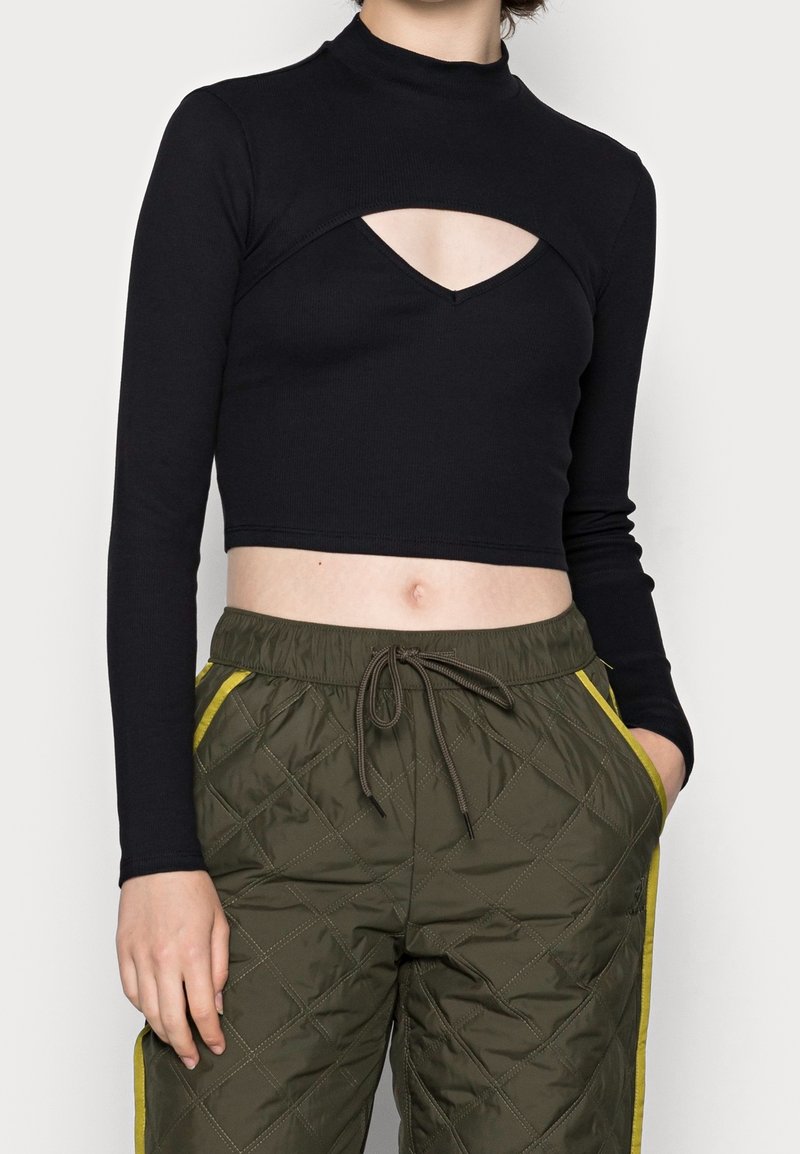 Haut noir court à manches longues avec un col montant et un détail découpé au niveau de la poitrine, associé à un pantalon de jogging matelassé vert olive avec des bandes jaunes.