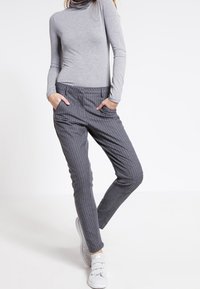 Grijs coltrui-jersey gecombineerd met grijze pantalon met streepdessin, voorzien van een elastische tailleband en zijzakken, gedragen met witte sneakers.