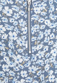 Top zip con motivo floreale blu e bianco, con una cerniera argentata al centro, tessuto testurizzato e motivi floreali sparsi.