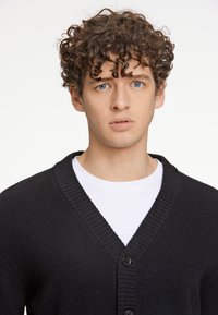 Svart kofta med v-ringning och ribbad krage, med tre knappar framtill. Bärs över en vit skjorta. Slät textur.