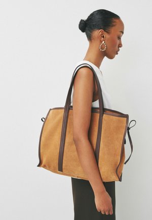 Woman with pulled-back hair wearing a sleeveless white top and large hoop earrings carries a tan suede tote bag with dark brown straps.