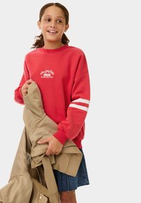 Felpa rossa con strisce bianche sulle maniche, logo ricamato sul petto, che tiene una giacca beige, abbinata a una gonna di jeans.