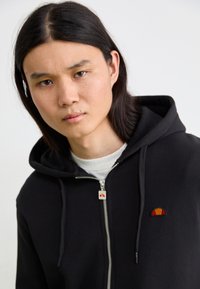 Schwarze Zip-Hoodie mit strukturierter Oberfläche, ausgestattet mit einem kleinen mehrfarbigen Logo auf der linken Brust. Kordeln und weiches Innenfutter sind sichtbar.