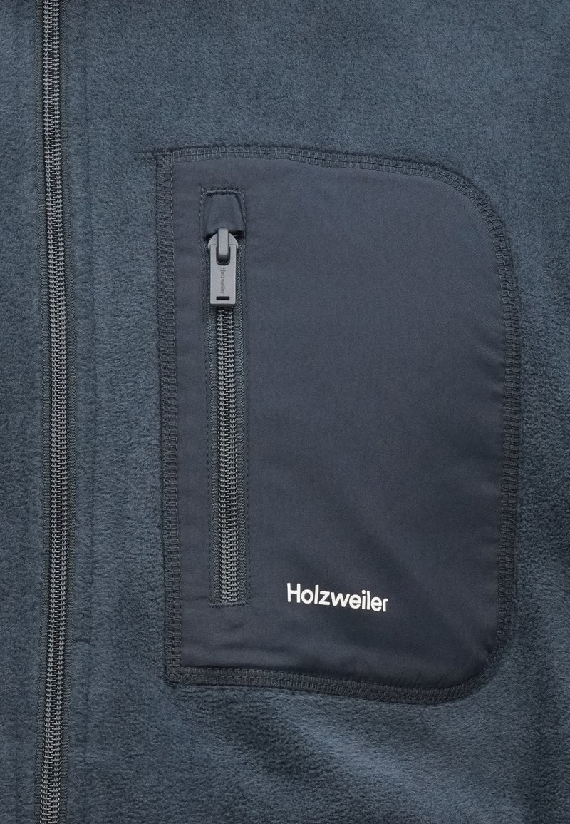 Veste en polaire bleu marine avec une poche zippée. Caractéristiques : tissu texturé, fermeture éclair centrale, et logo "Holzweiler" en blanc sur la poche.